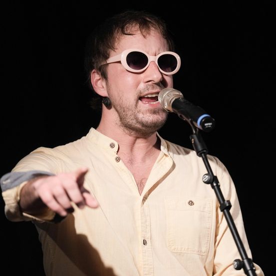 Ein Mann in einem hellgelben Hemd und rosa Sonnenbrille singt in ein Mikrofon. Er zeigt mit der linken Hand.