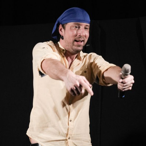 Ein Mann mit blauer Bandana und gelbem Hemd steht auf der Bühne, hält ein Mikrofon in der Hand und zeigt mit dem Finger nach vorne.