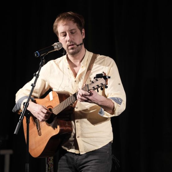 Ein Mann spielt Gitarre auf der Bühne, hält sie nah an sein Gesicht. Er trägt ein gelbes Hemd und spielt in der Nähe eines Mikrofons auf einem Ständer.