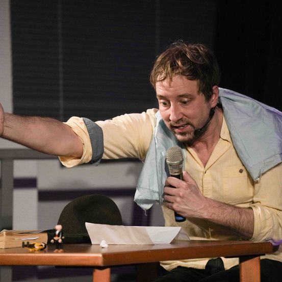 Ein Mann sitzt an einem Tisch und hält ein Mikrofon in der Hand. Er trägt ein gelbes Hemd und eine blaue Decke über den Schultern.