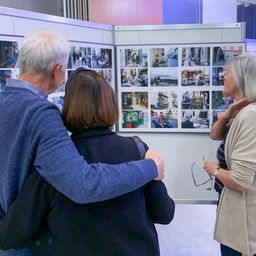 Ein älteres Paar und eine andere Frau stehen vor einer Fotoausstellung und betrachten die Bilder. Der Mann hat seinen Arm um die Schulter der Frau gelegt.