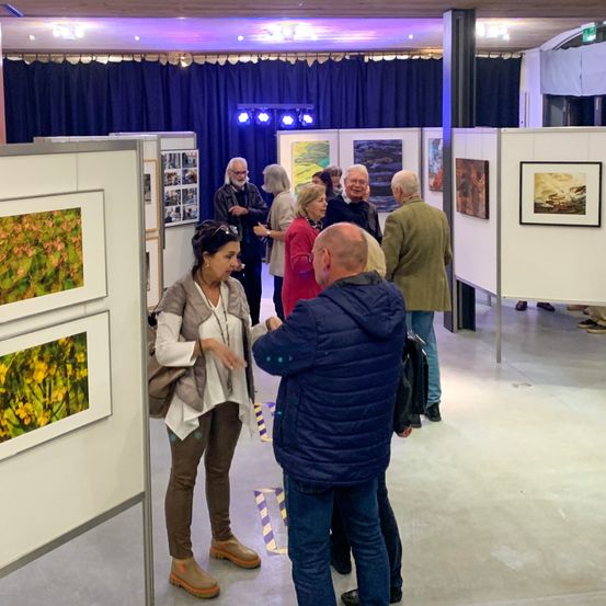 Eine Gruppe von Menschen versammelt sich in einer überdachten Galerie und bewundert verschiedene gerahmte Gemälde an den Wänden. Einige Leute unterhalten sich, während andere die Kunstwerke betrachten.