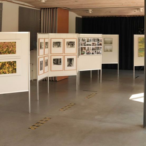 Ein Innenbereich zeigt mehrere Fotocollagen auf weißen Ständern. Einige Fotos sind gerahmt, andere sind Collagen. Der Boden ist aus Beton, und auf dem Boden befinden sich Warnbänder.