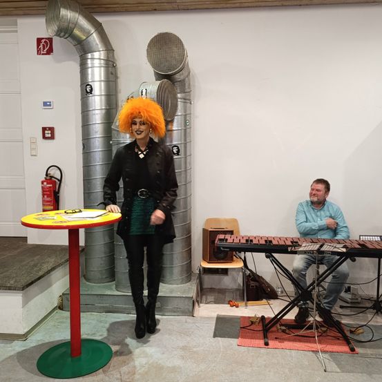 Eine Frau mit orangefarbenem Haar steht neben einem rot-gelben Tisch, während ein Mann am Klavier sitzt. Im Hintergrund befinden sich Metallrohre und ein Feuerlöscher.