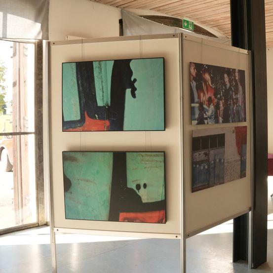 Ein Metallständer mit vier ausgestellten Gemälden in einem Raum. Das Gemälde oben links ist dunkelgrün und schwarz. Das Gemälde oben rechts zeigt Menschen. Das Gemälde unten links ist dunkelgrün mit rot. Das Gemälde unten rechts ist weiß mit rot.