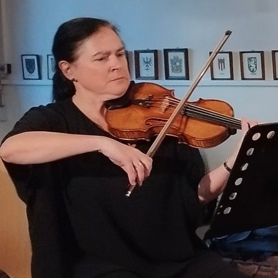 Eine Frau spielt Geige in einem Raum mit gerahmten Bildern an der Wand. Sie hält die Geige mit der linken Hand und den Bogen mit der rechten.