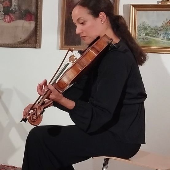 Eine Frau sitzt und spielt Geige. Sie trägt ein schwarzes Kleid, und ihr Haar ist zu einem Pferdeschwanz gebunden. Zwei gerahmte Gemälde schmücken die Wand hinter ihr.