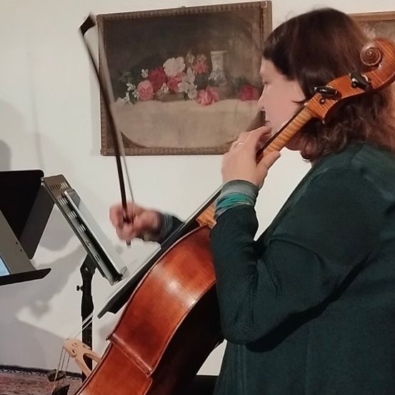 Eine Frau spielt Cello im Sitzen, neben einem Notenständer. Sie ist in Grün gekleidet, und die Szene spielt drinnen.