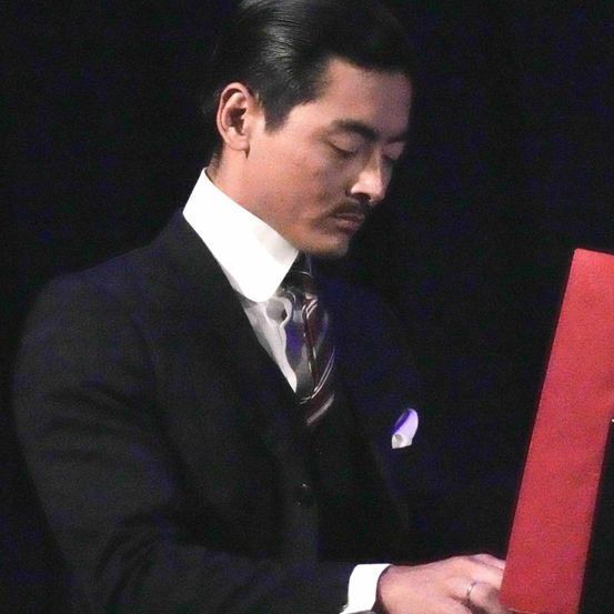 Ein Mann in einem schwarzen Anzug hält eine rote Mappe und schaut nach unten, vielleicht bei einer Preisverleihung. Er hat einen ernsten Ausdruck und einen weißen Kragen.