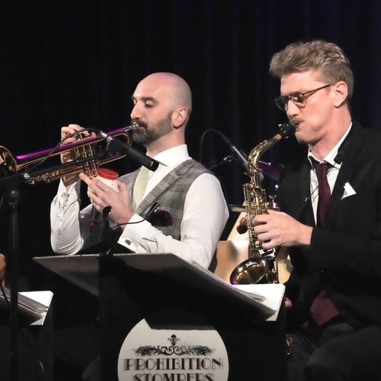 Zwei Männer spielen auf der Bühne mit Trompete und Saxophon vor einem schwarzen Hintergrund mit einem Prohibition Stompers Logo. Ein Mann hält einen roten Becher, und beide tragen Anzüge und Krawatten.