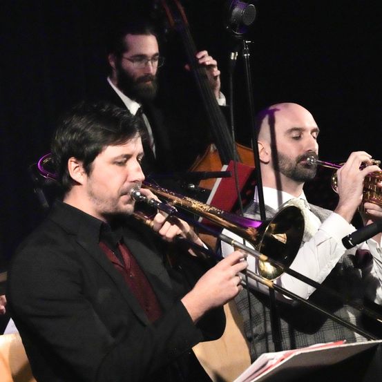 Drei Männer spielen auf der Bühne Musikinstrumente, einer spielt Trompete, der andere Posaune und der dritte Cello. Die Bühne ist schwach beleuchtet.