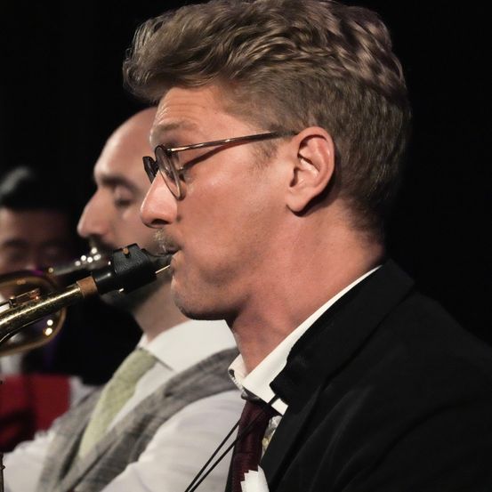Ein Mann mit Brille und Anzug spielt die Saxophon auf der Bühne, während ein Trompeter hinter ihm steht.