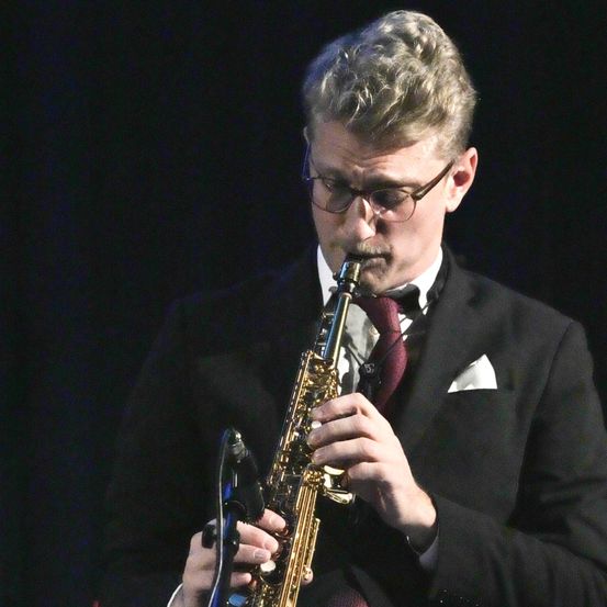 Ein Mann in einem schwarzen Anzug und mit Brille spielt eine Saxophon auf der Bühne mit einem Mikrofon vor sich.