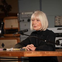 Eine ältere Frau mit kurzen weißen Haaren sitzt an einem Tisch und spricht in ein Mikrofon. Sie trägt ein schwarzes Rollkragenshirt. Ein Buch und eine Lampe liegen auf dem Tisch.