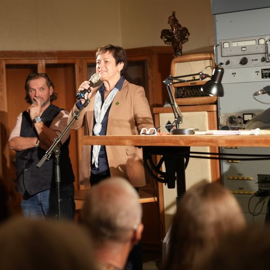 Eine Frau spricht in ein Mikrofon auf der Bühne, begleitet von einem Mann, der hinter ihr steht. Ein Publikum sitzt vor ihnen und ein Schreibtisch mit einer Lampe steht auf der Bühne.