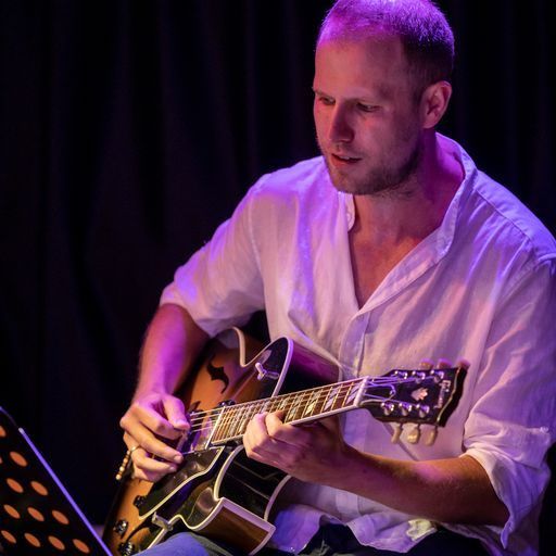 Ein Mann spielt Gitarre unter lila Bühnenbeleuchtung. Er trägt ein weißes Hemd und Jeans und konzentriert sich auf sein Instrument, während ein unscharfer schwarzer Vorhang im Hintergrund zu sehen ist.