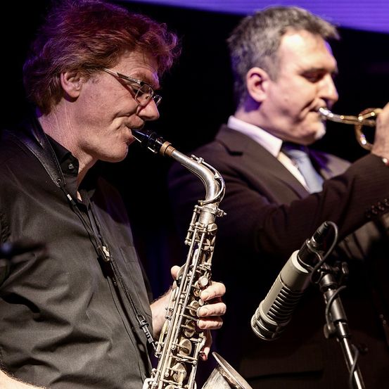Zwei Männer spielen auf der Bühne, einer spielt Saxophon und der andere Trompete, beide stehen vor Mikrofonen.