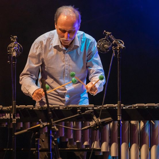 Ein älterer Mann spielt ein Xylophon auf der Bühne, beleuchtet von blauen Lichtern, mit Mikrofonen und Instrumenten um ihn herum.