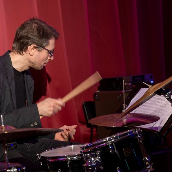 Ein Mann mit Brille spielt auf der Bühne Schlagzeug mit einem roten Vorhang dahinter.