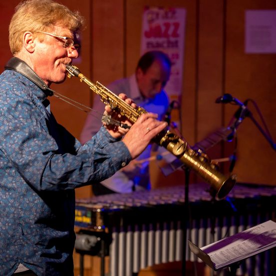 Ein älterer Mann mit Brille spielt Saxophon bei einem Jazz-Konzert. Hinter ihm spielt ein anderer Mann Marimba.