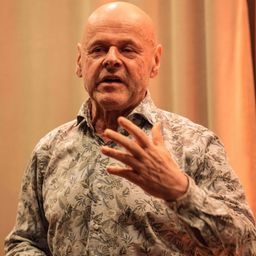 Ein älterer Mann in einem Blumenhemd spricht mit Handgesten, sieht ernst aus, mit einem unscharfen Vorhang im Hintergrund.