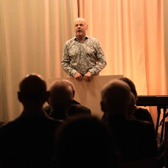 Ein älterer Mann spricht an einem Rednerpult und wendet sich an eine Gruppe von sitzenden Zuhörern. Die Bühne ist mit warmen, pfirsichfarbenen Vorhängen geschmückt.