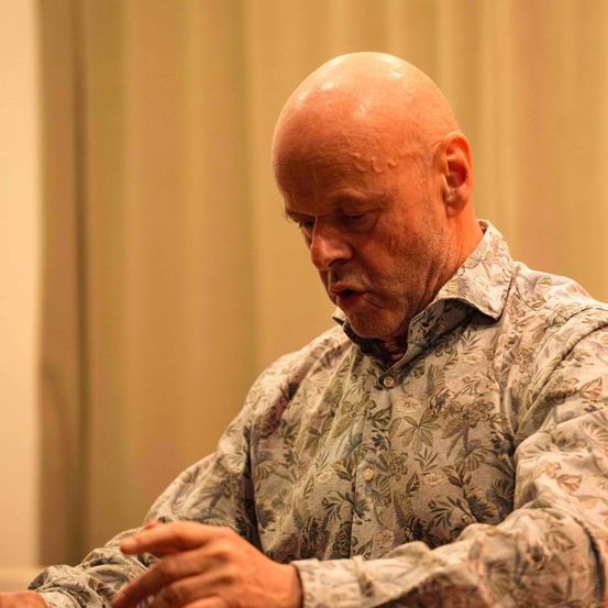 Ein älterer kahlköpfiger Mann in einem Blumenhemd sitzt am Tisch und schaut mit geschlossenen Augen nach unten.