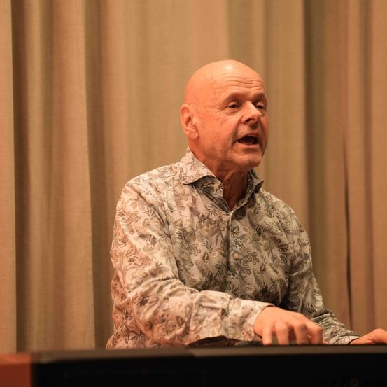 Ein älterer Mann mit kahlem Kopf sitzt an einem Schreibtisch, wahrscheinlich in einem Konferenzraum, und scheint zu sprechen. Er trägt ein Hemd mit Blumenmuster.