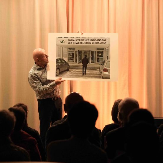 Ein Mann spricht vor einem Publikum und hält ein Poster hoch, das ein Gebäude und eine Person zeigt, die weggeht.