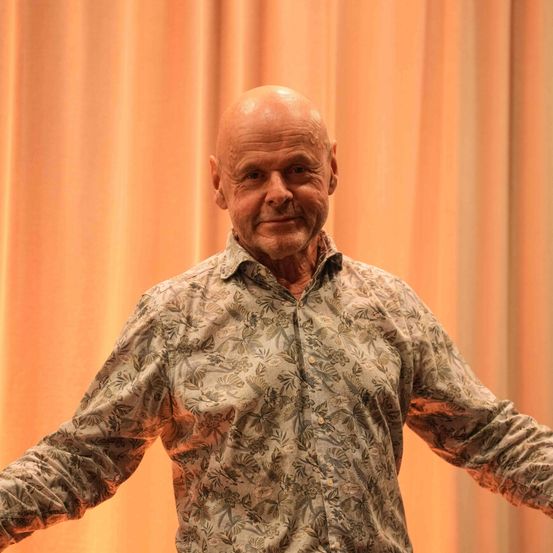 Ein älterer Mann steht mit ausgestreckten Armen auf einer Bühne. Er trägt ein Blumenhemd und schwarze Hose. Hinter ihm ist ein orangefarbener Vorhang.