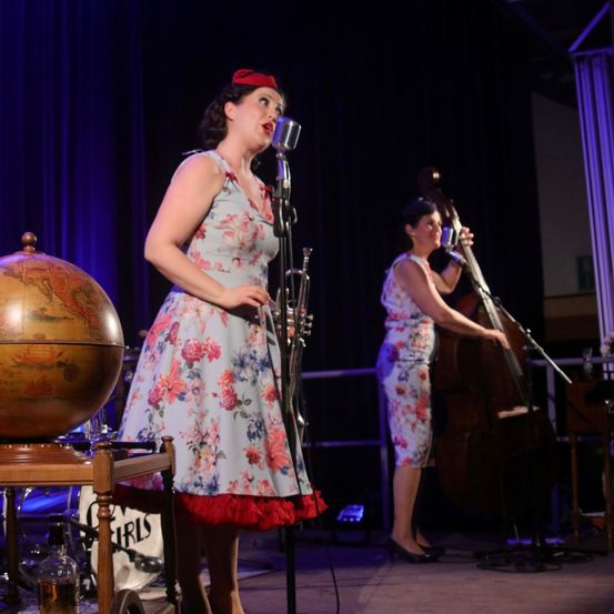 Eine Frau singt in ein Mikrofon auf der Bühne, mit einem Globus und einer Trompete in der Nähe. Eine andere Frau spielt Cello. Die Bühne ist mit blauem Licht beleuchtet.