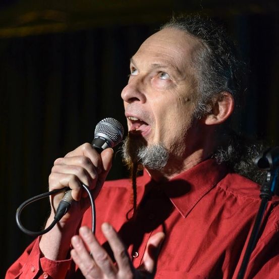 Ein Mann mit langem Bart singt in ein Mikrofon, sein Gesicht von Bühnenlichtern beleuchtet, das Mikrofon mit beiden Händen haltend.