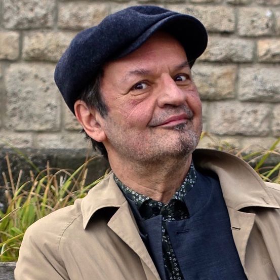 Ein Mann mit Bart und Schnurrbart, der einen schwarzen Hut und eine beige Jacke trägt, lächelt und sitzt vor einer Ziegelmauer.