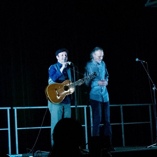 Zwei Männer treten auf der Bühne auf. Einer hält eine Gitarre, während der andere in ein Mikrofon singt. Die Bühne ist dunkel mit einer weißen Brüstung.