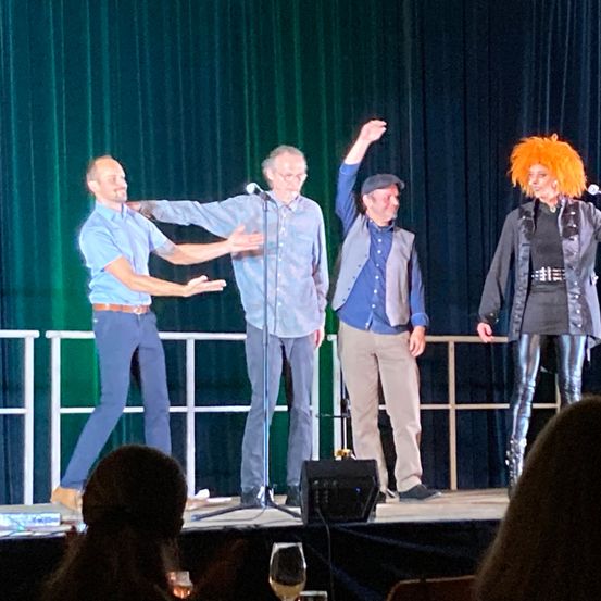 Vier Personen stehen auf der Bühne, zwei von ihnen sprechen in Mikrofone. Eine Frau mit orangefarbenem Haar steht neben einem Mann in Lederhosen. Hinter ihnen deutet ein Mann mit erhobenen Händen.