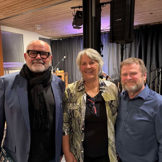 Drei Personen stehen nah beieinander und lächeln in einem Raum. Der Raum hat Vorhänge, Lautsprecher und eine Holzdecke. Der Mann auf der linken Seite trägt einen Schal und eine Brille. Die Frau in der Mitte hat eine Halskette. Der Mann auf der rechten Seite trägt ein blaues Hemd.