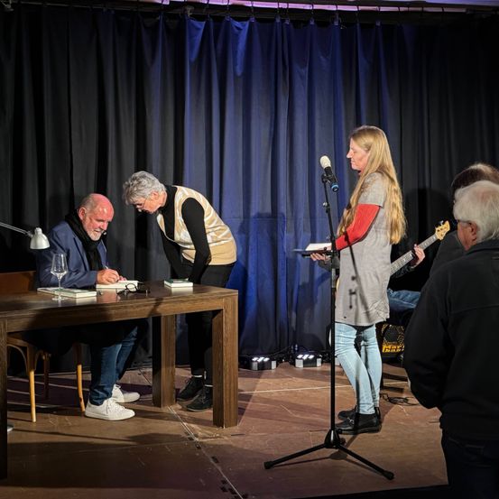 Ein Mann sitzt an einem Tisch und signiert Bücher, während eine Frau von einem Tablet liest. Hinter ihnen spielt ein Mann Gitarre. Im Hintergrund hängt ein blauer Vorhang.