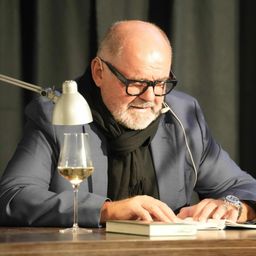 Ein älterer Mann mit Brille und Schal liest ein Buch mit einem Glas Wein auf dem Tisch vor ihm.