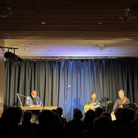 Drei Musiker treten auf der Bühne in einem schwach beleuchteten Raum mit blauem Vorhang im Hintergrund auf. Ein Mann liest sitzend aus einem Buch, während die anderen zwei Gitarre spielen. Das Publikum sitzt vor ihnen.