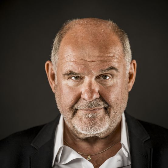 Ein Nahporträt eines älteren Mannes mit weißem Bart und Schnurrbart, der einen schwarzen Anzug und ein weißes Hemd trägt, vor einem dunklen Hintergrund.