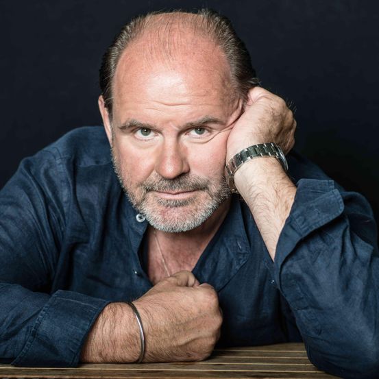 Einer mittleren Alters mit Bart, der ein dunkelblaues Hemd trägt, sitzt mit der Hand am Kopf und blickt zur Seite.