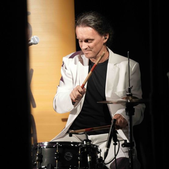 Ein Mann spielt auf der Bühne Schlagzeug, trägt einen weißen Anzug und ein schwarzes Hemd. Er konzentriert sich auf das Schlagzeug, hält Trommelstöcke in der Hand, und ein Mikrofon ist in der Nähe.