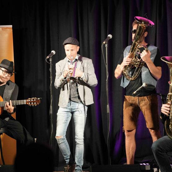 Eine Band spielt auf einer Bühne mit schwarzem Vorhang. Der Trompeter steht in der Mitte mit einem Mikrofon. Auf seiner linken Seite sitzt ein Gitarrist auf einem Stuhl. Auf seiner rechten Seite steht ein Tubist. Eine weitere Person ist teilweise sichtbar auf der rechten Seite und spielt Saxophon.