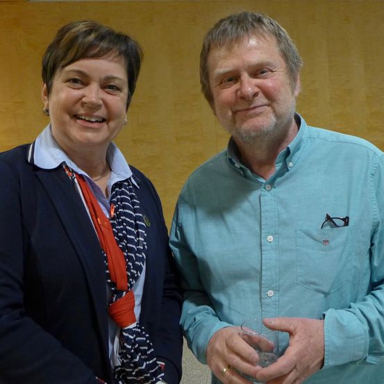 Ein Mann und eine Frau stehen eng beieinander und lächeln, wahrscheinlich bei einer Veranstaltung. Der Mann hält ein Glas. Hinter ihnen ist eine Holzwand.