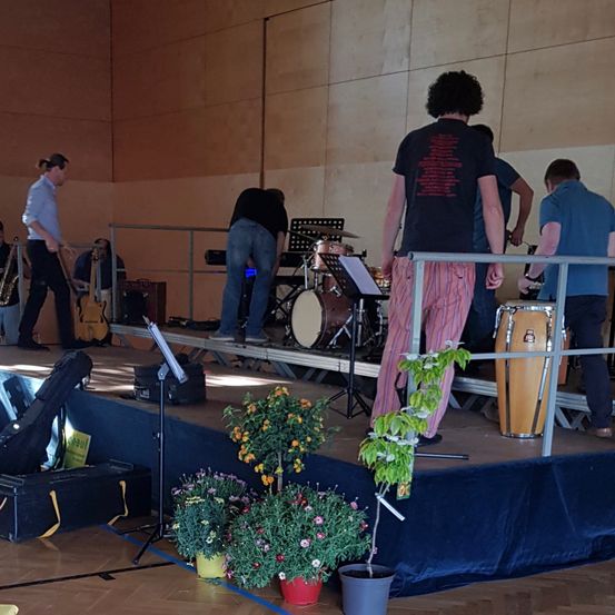 Eine Gruppe von Musikern bereitet sich auf einer Bühne mit verschiedenen Instrumenten vor, darunter eine Saxophon, eine Gitarre und Trommeln. Topfpflanzen und Notenständer sind um die Bühne herum aufgestellt. Die Musiker tragen Freizeitkleidung.