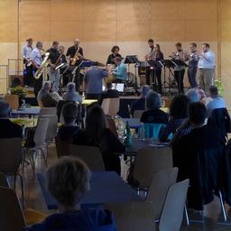 Eine Gruppe Musiker tritt auf einer Bühne in einem Saal mit Holzwänden auf. Das Publikum sitzt auf Stühlen vor Tischen. Einige stehen in der Nähe der Bühne, andere beobachten von weiter weg.