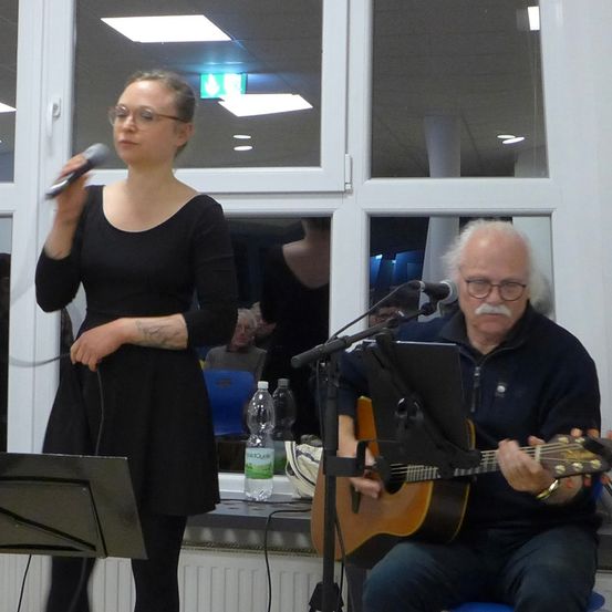 Eine Frau singt in ein Mikrofon, während ein Mann vor einem Glasfenster die Gitarre spielt.