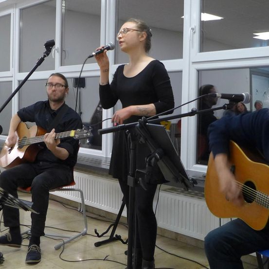 Eine Frau singt in ein Mikrofon, während zwei Männer Gitarre spielen. Der Mann auf der linken Seite sitzt, der Mann auf der rechten Seite steht. Hinter ihnen befinden sich Fenster und ein Heizkörper.