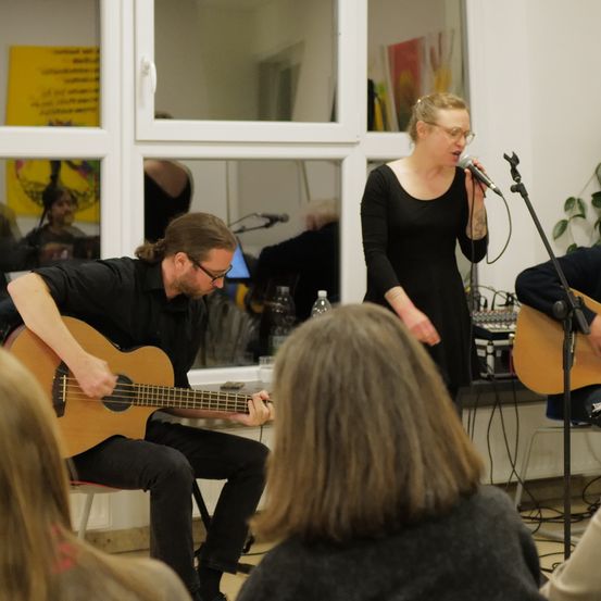Eine Frau singt, während sie an einem Mikrofon vor einem Fenster steht. Ein Mann spielt Gitarre vor ihr. Eine weitere Person spielt Gitarre rechts. Ein Publikum schaut zu.