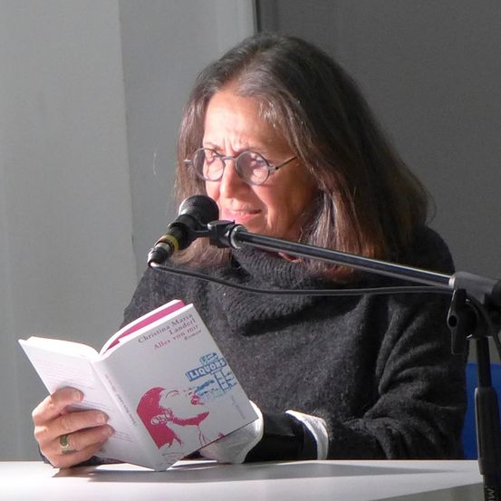Eine Frau mit Brille sitzt an einem Tisch und liest ein Buch mit dem Titel 'Christina Maria Landler'. Sie trägt einen Pullover und hat ein Mikrofon vor sich.